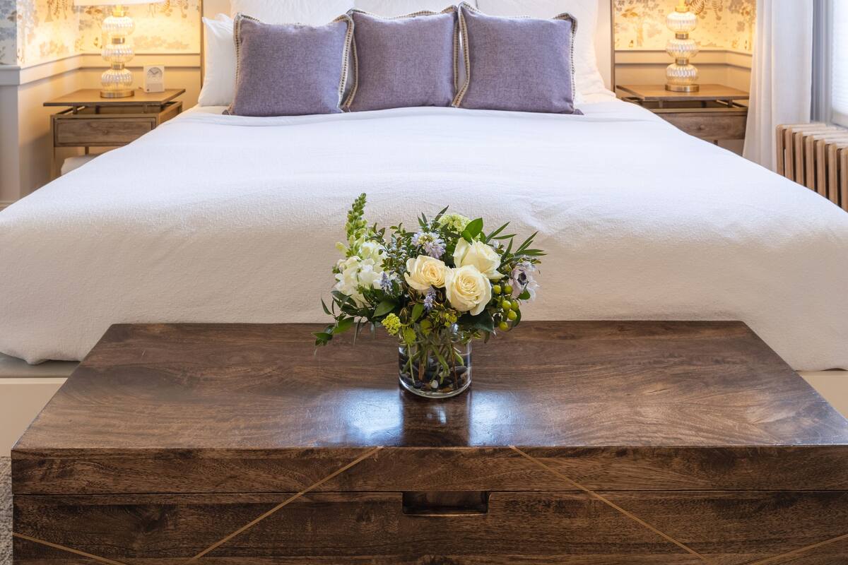 A bedroom with a large bed, two bedside tables with lamps, a wooden coffee table with a vase of flowers, and patterned wallpaper.