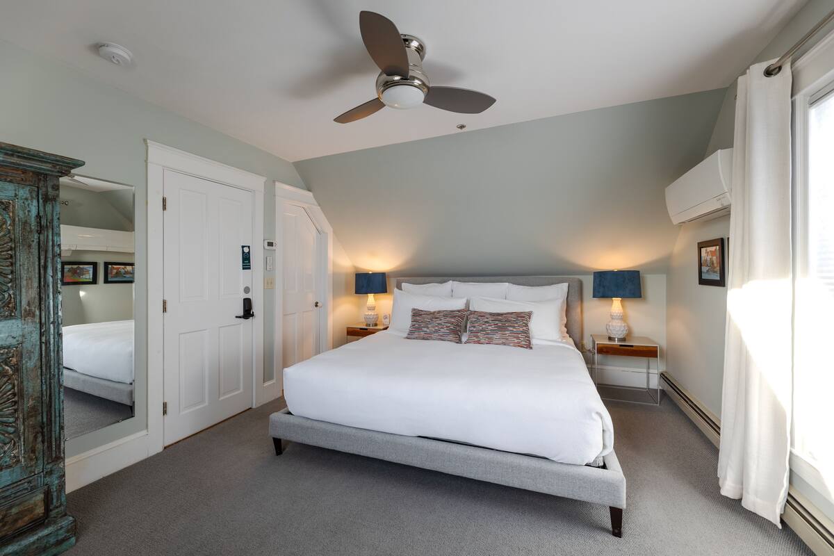 A bedroom with a large bed, a ceiling fan, bedside lamps, and a view of the outside.