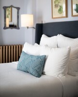 Una cama bien hecha con almohadillas blancas y azules estampadas, un cabecero oscuro y una lámpara fijada a la pared.