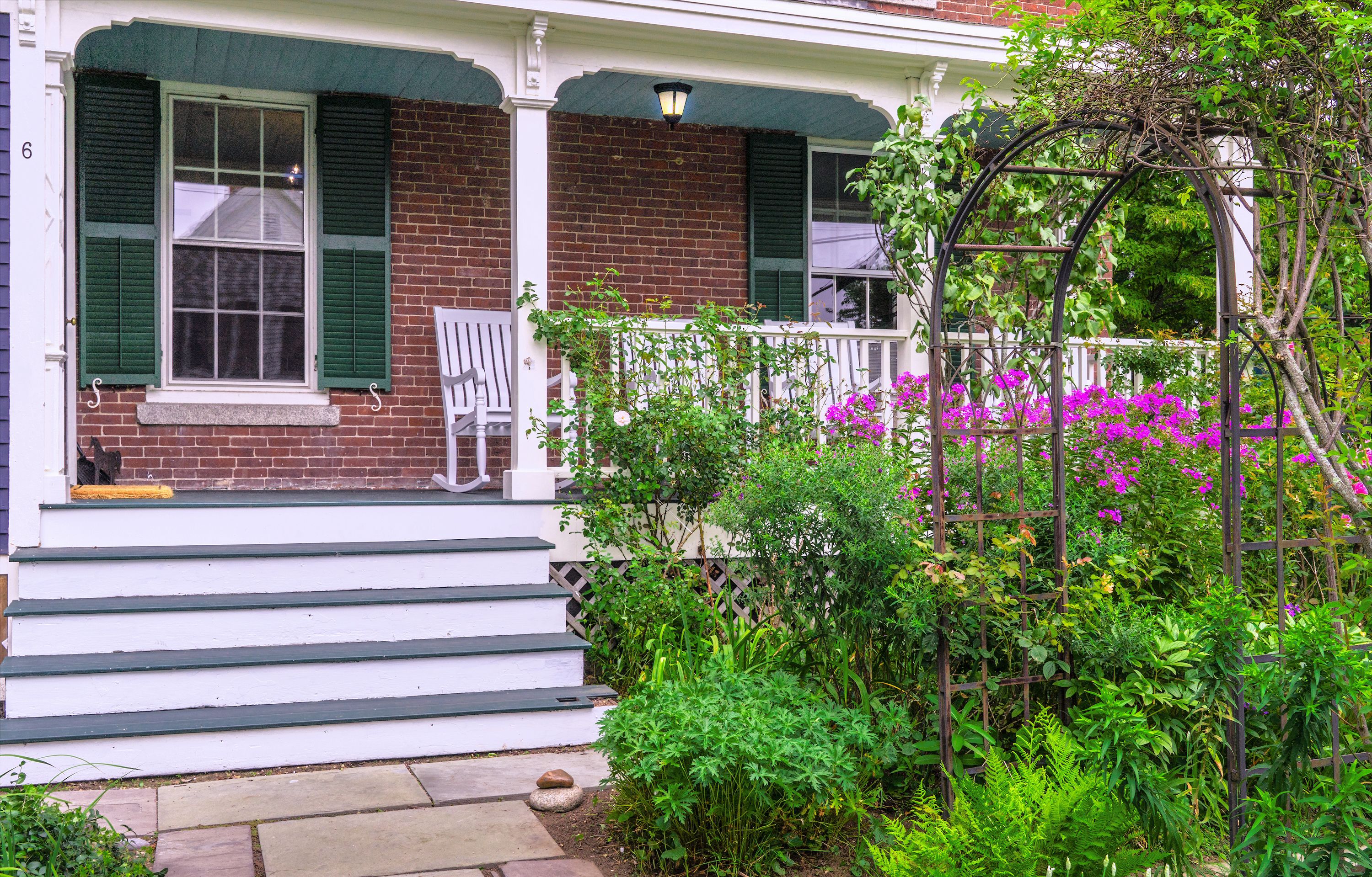 A brick house with a porch, green shutters, and a garden with pink flowers.