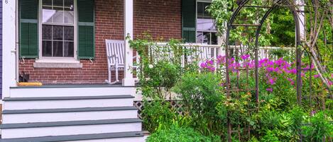 A brick house with a porch, green shutters, and a garden with pink flowers.