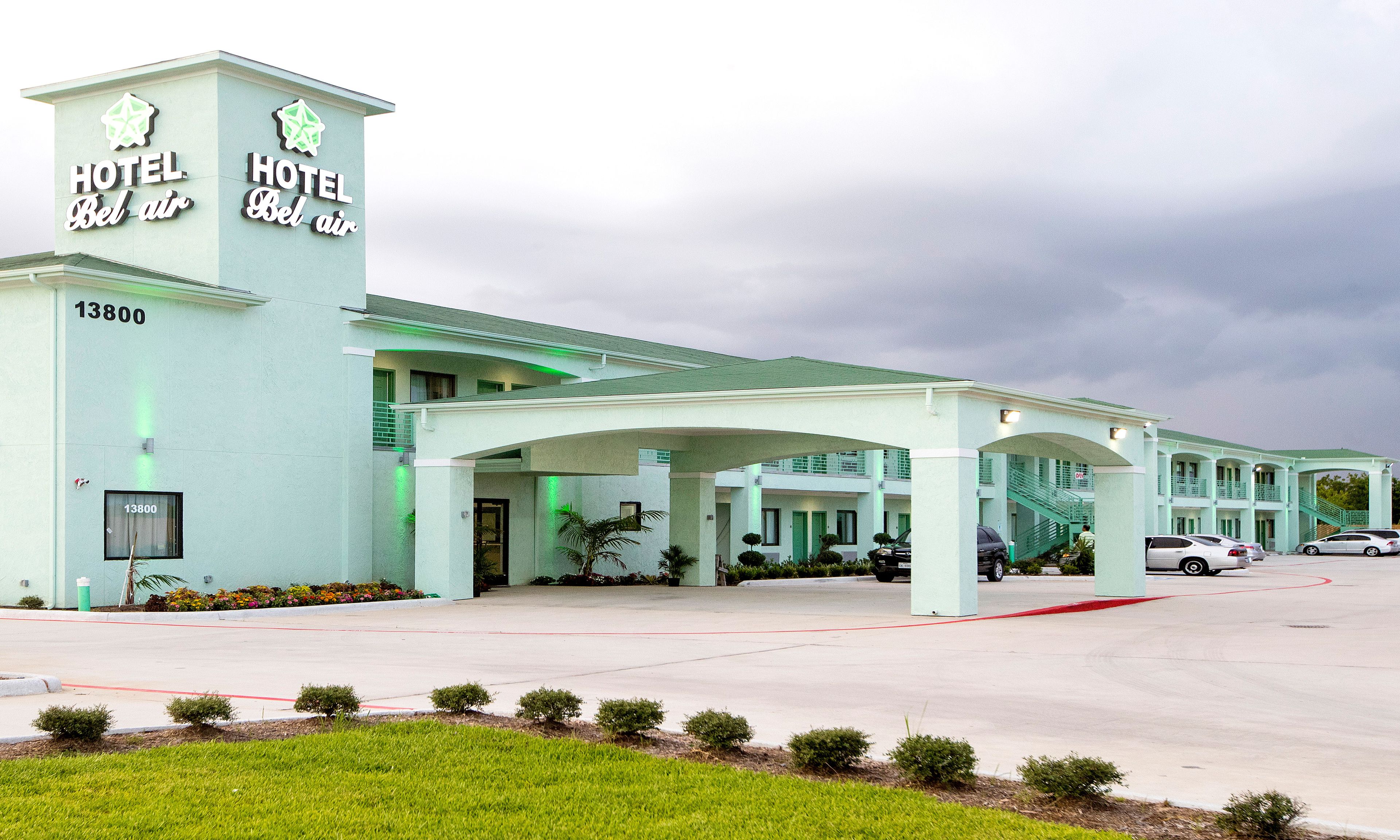 Un hotel con fachada verde claro, un letrero llamativo que dice "HOTEL Bel Air" y un estacionamiento con varios autos.