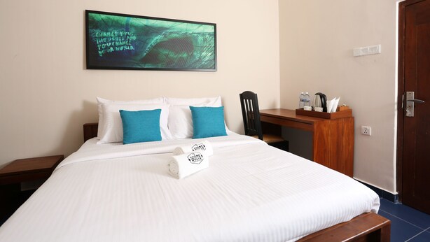 A neatly made bed with white and teal bedding, a wall-mounted abstract painting, a wooden desk with a chair, and a door. - Boots Travelers Lodge (Negombo)