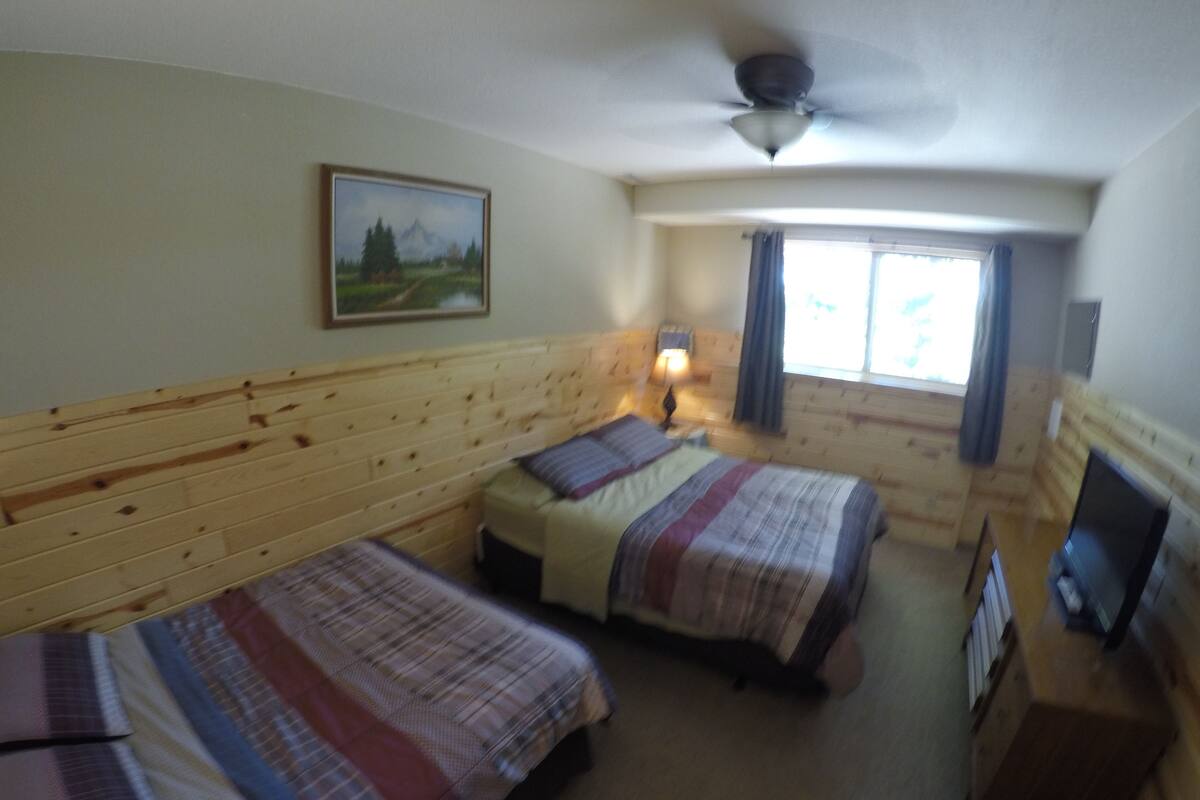 A room with two beds, a ceiling fan, a painting on the wall, a window with curtains, and a television on a wooden cabinet.