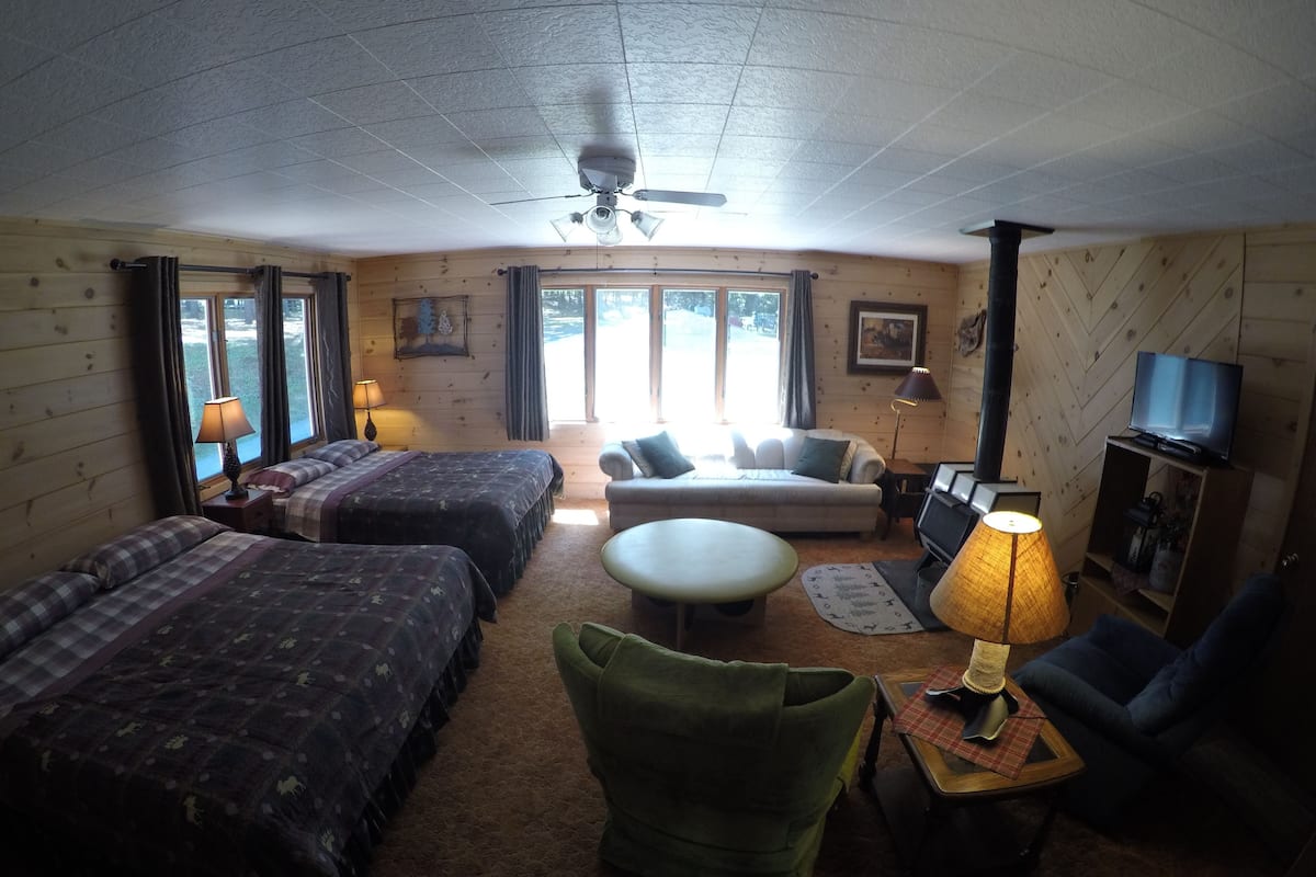 A cozy cabin-style room with two beds, a sofa, a green armchair, a TV, and a fireplace.