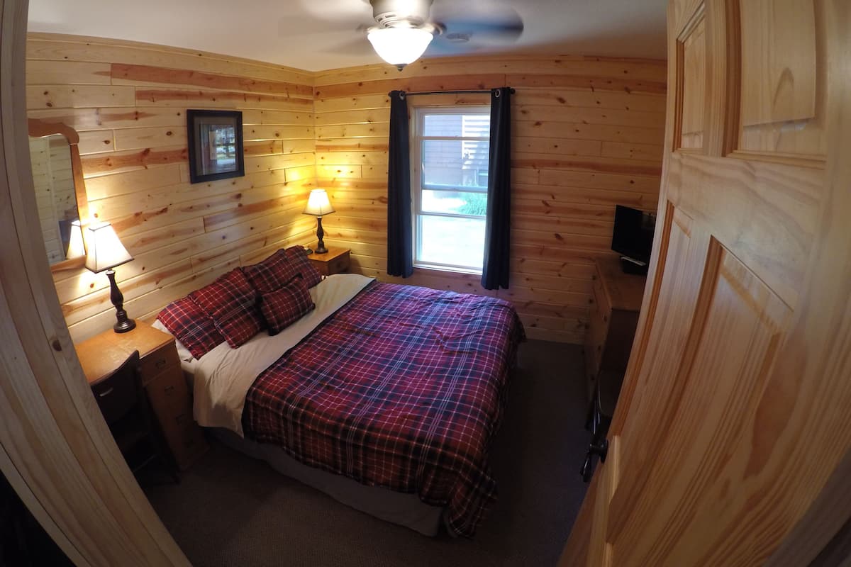 A cozy room with wooden walls, a bed with plaid bedding, two bedside lamps, a ceiling fan, and a window with curtains.