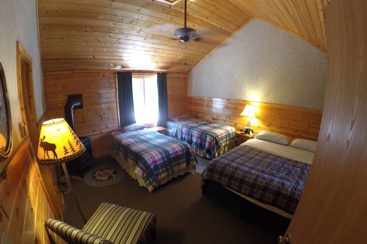 A cabin-style room with two beds, a wooden ceiling, a lamp with a deer silhouette, and a small table.
