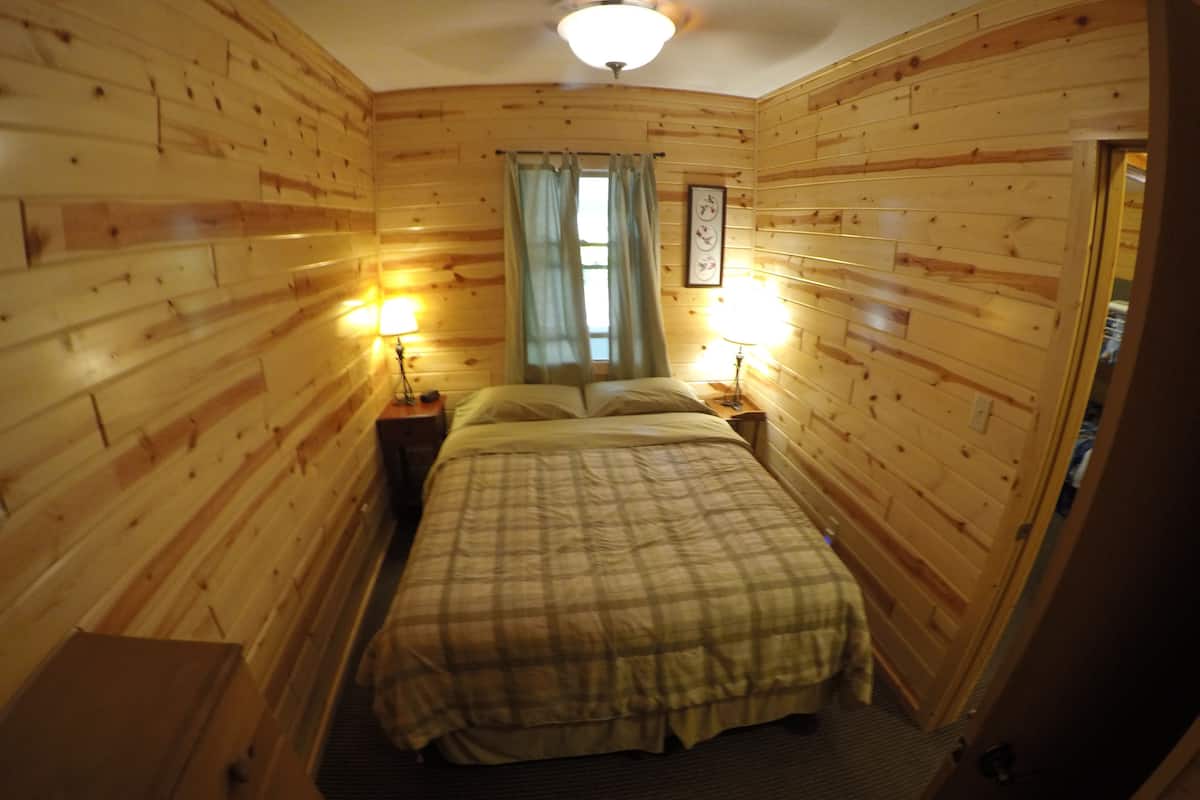 A wooden-paneled room with a bed, two bedside lamps, a window with curtains, and a door.