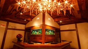 A wooden reception desk with a chandelier above, wooden beams on the ceiling, and two aquariums on the wall. - Estrada Real Palace Hotel (Brumadinho)