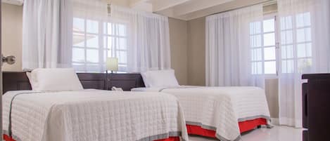 A hotel room with two beds, each with white bedding and red skirts, a wooden headboard, and a window with sheer curtains.