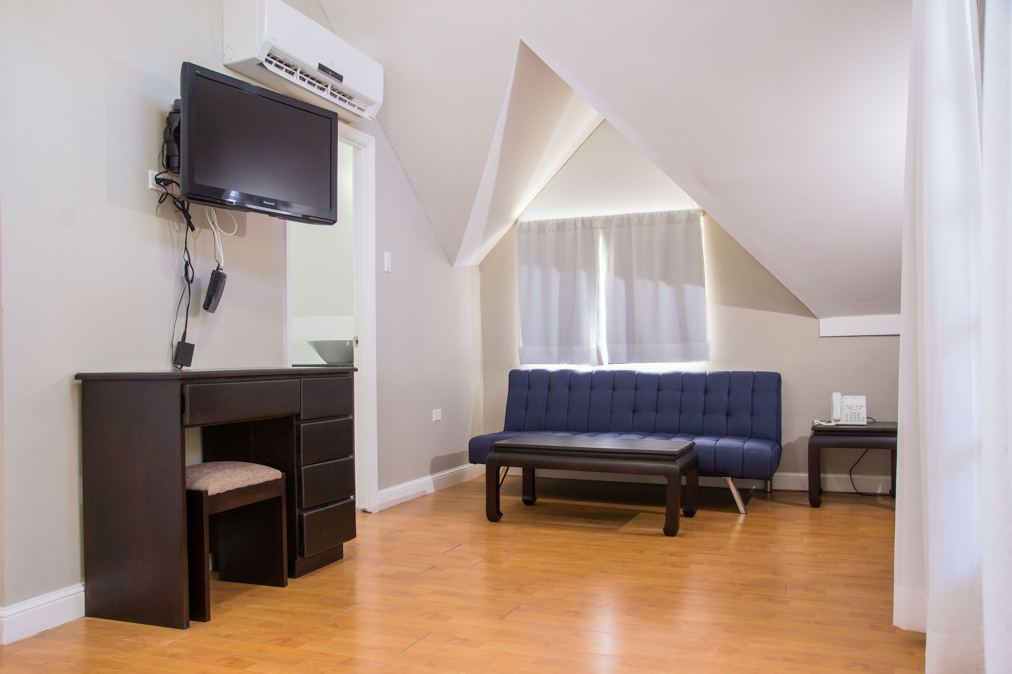 A room with a dark wooden desk, a blue sofa, a small table, and a TV mounted on the wall.