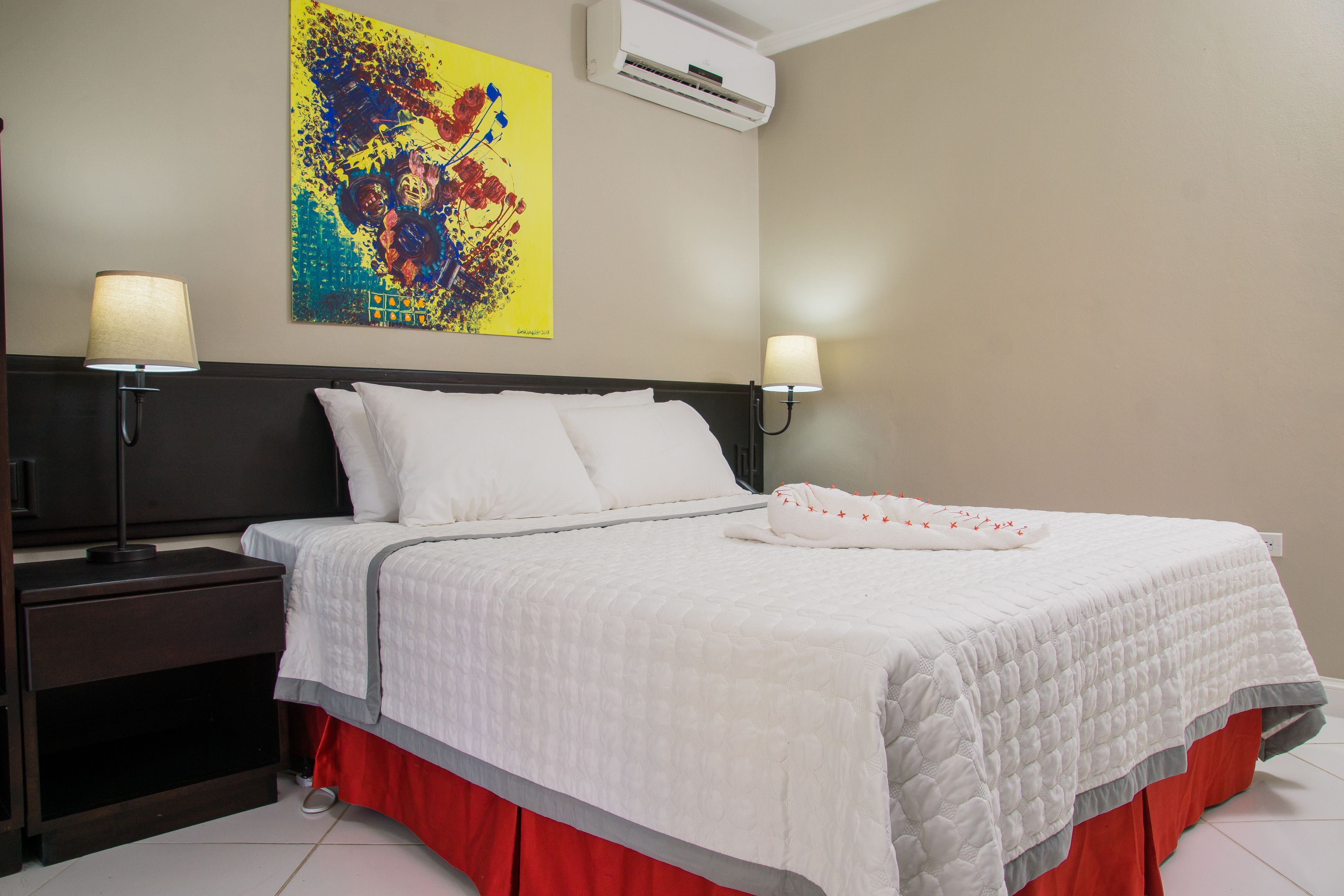 Habitación de hotel con una cama grande, dos lámparas de noche, aire acondicionado de pared y un cuadro abstracto colorido.