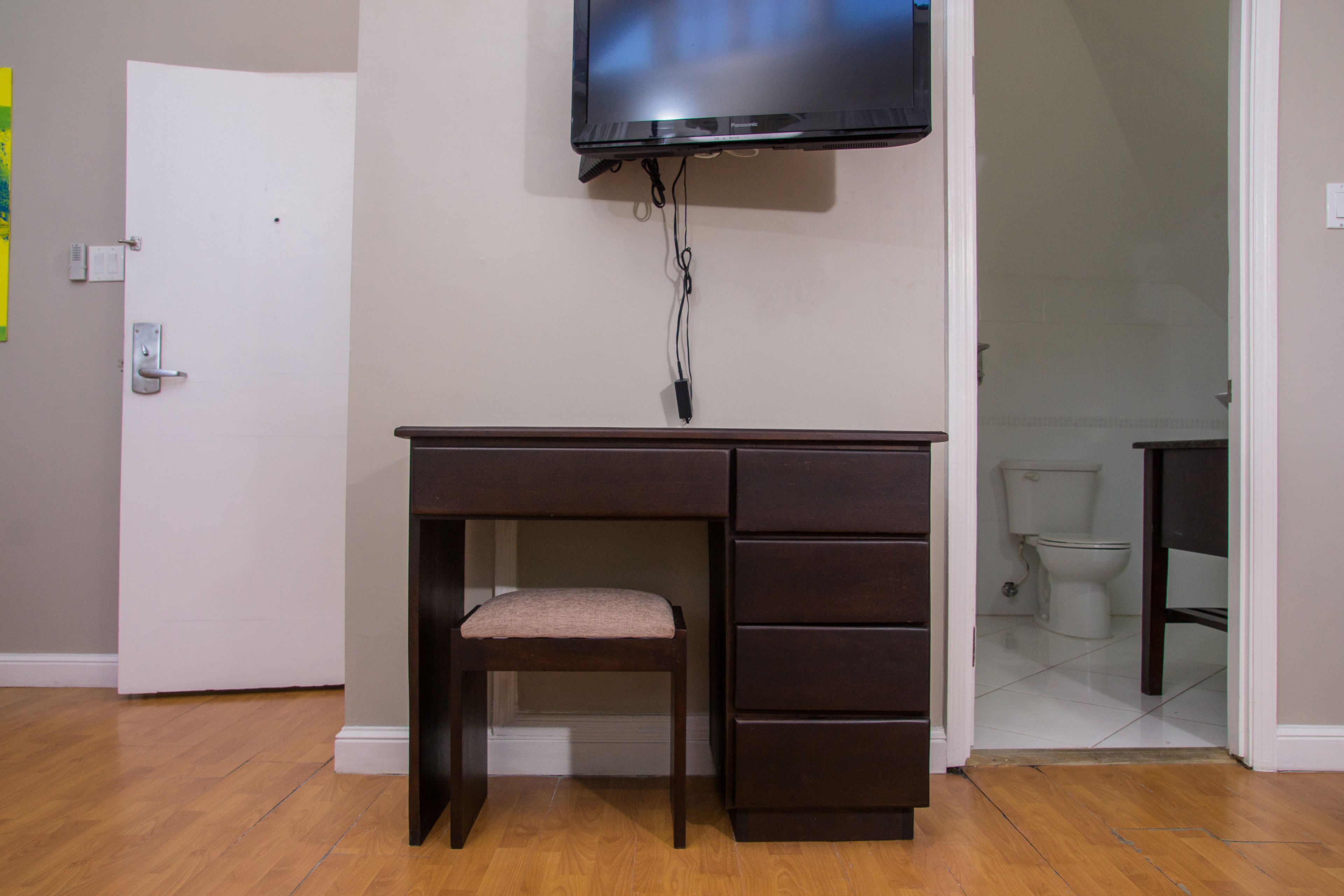 A room with a wooden desk, a chair, a television mounted on the wall, and a door leading to a bathroom.