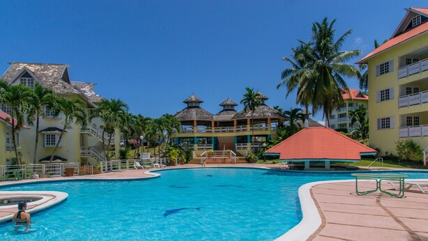 A resort pool area with a clear blue pool, palm trees, and resort buildings with red roofs.
