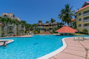 A resort pool area with a clear blue pool, palm trees, and resort buildings with red roofs.