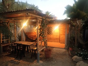 A rustic outdoor seating area with a wooden table and chairs, a hammock, and a wooden structure providing shade.