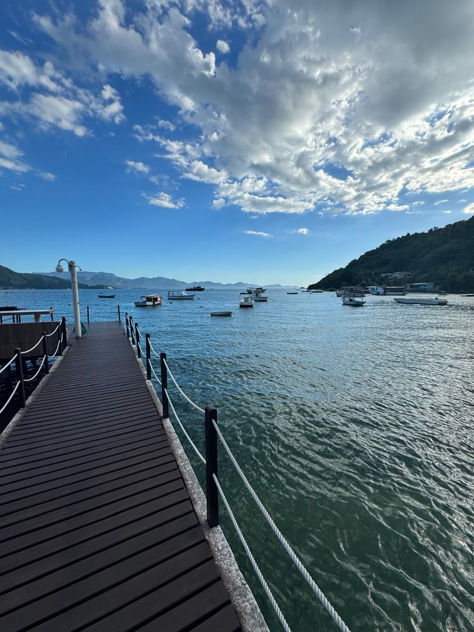 Une jetée en bois s’étendant sur une étendue d’eau calme, avec des bateaux, un ciel dégagé et un paysage vallonné à l’horizon.