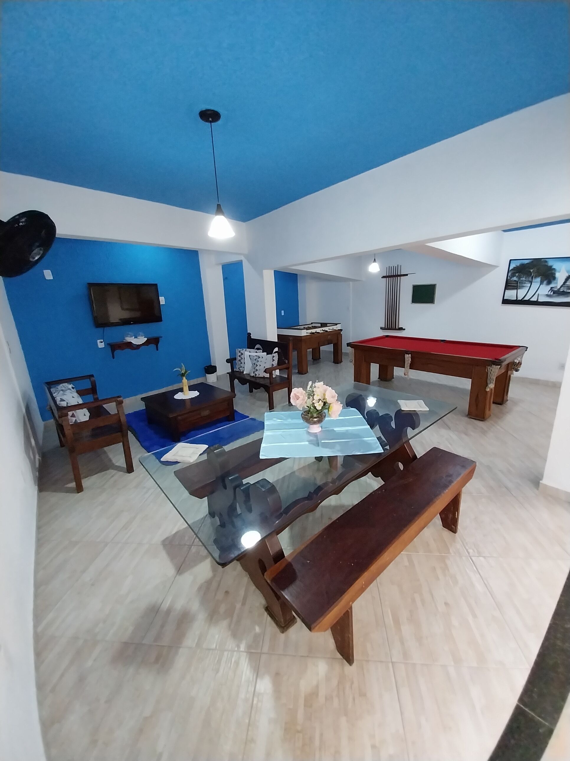 A room with a glass-top pool table, wooden benches, a sofa, a TV mounted on the wall, and a ceiling fan.