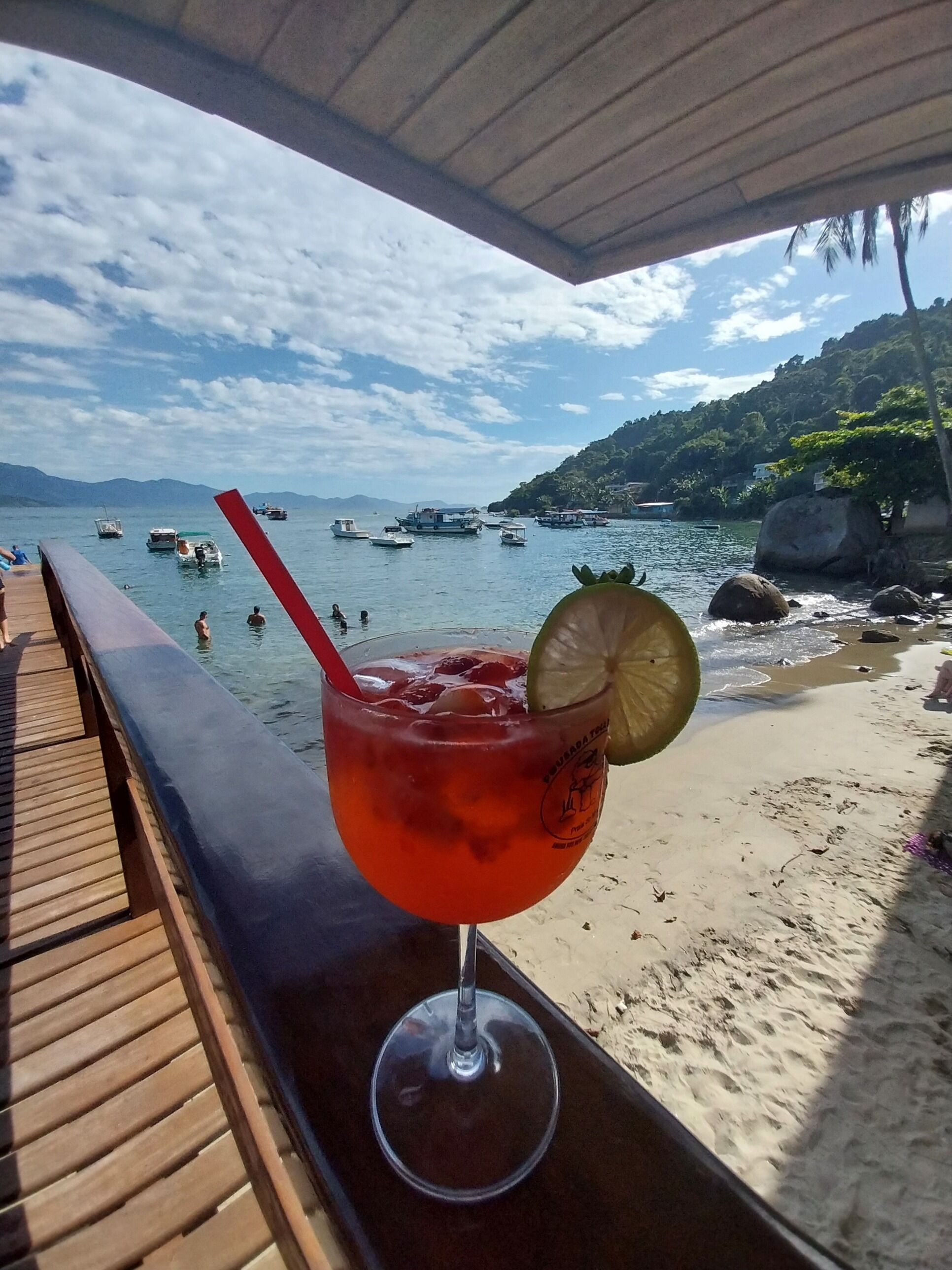 A beach view with boats on the water and a drink on a wooden bench.