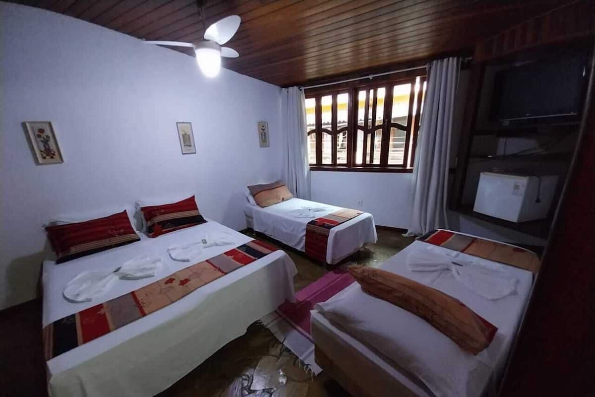 A room with two single beds, a ceiling fan, a window with curtains, and a television on a shelf.