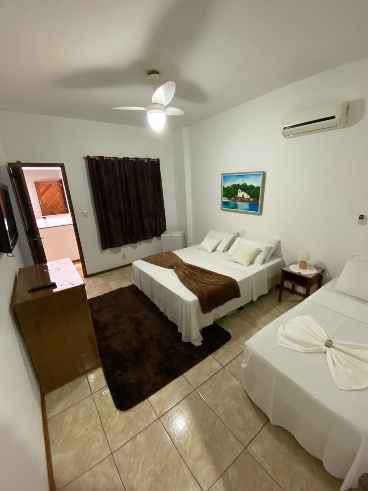 A hotel room with a bed, a desk, a chair, a small table, a nightstand, a picture on the wall, and an air conditioning unit.