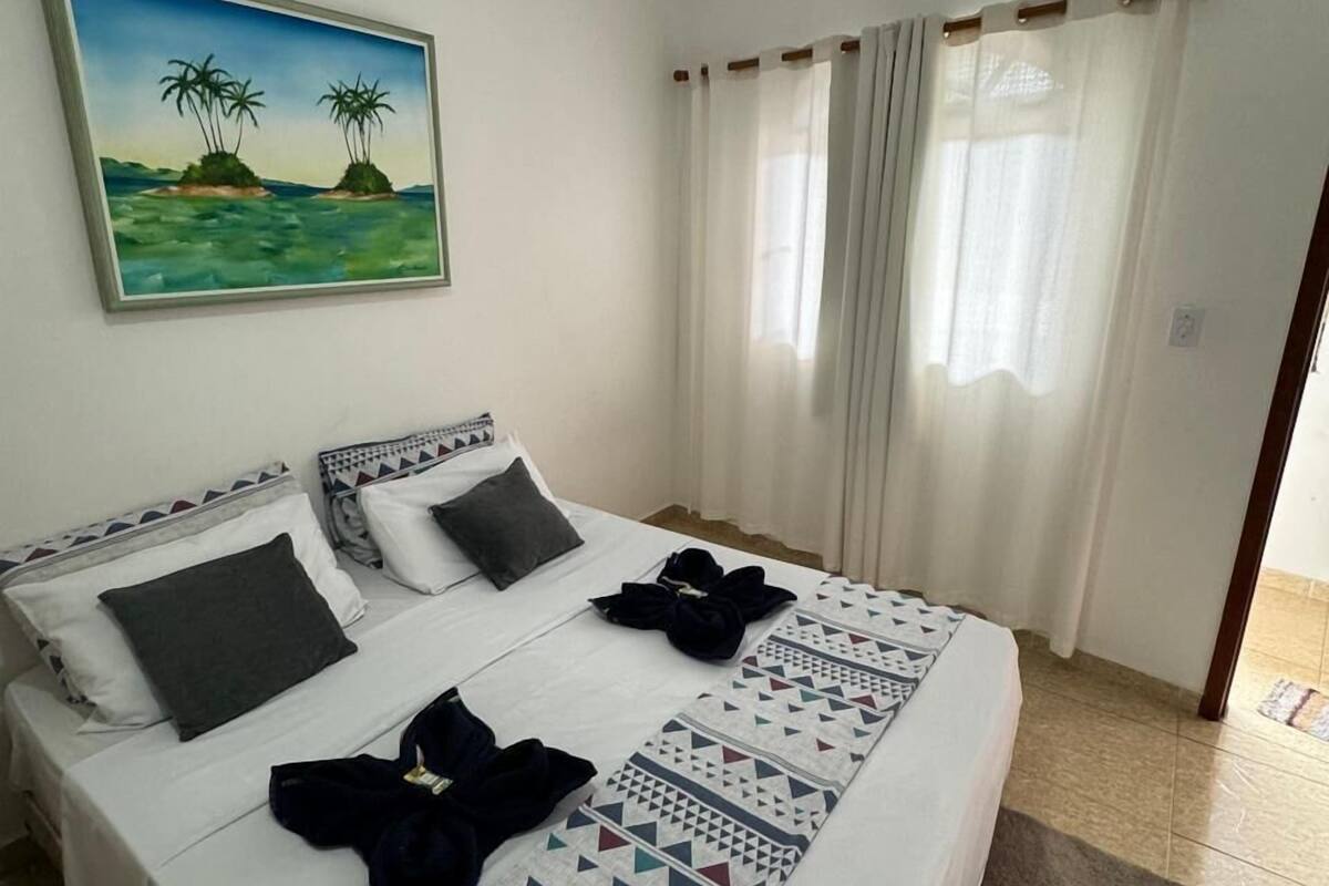 A hotel room with a bed, two pillows, a blanket, a painting, and a window with curtains.