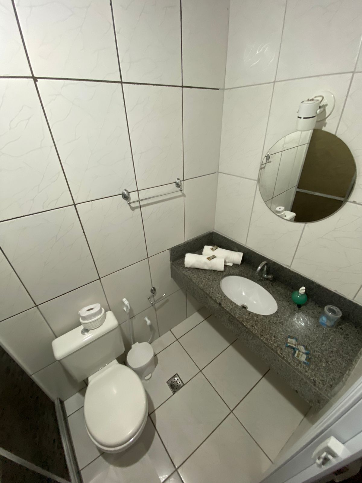 A bathroom with white tiled walls, a white toilet, a sink with a granite countertop, and a mirror.