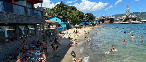 A beach with people swimming and sunbathing, a green surfboard, and a building with balconies.