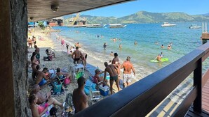 A beach scene with people sunbathing and swimming, a pier extending into the water, and boats in the background. - Pousada Toca do Cambú (Angra dos Reis)