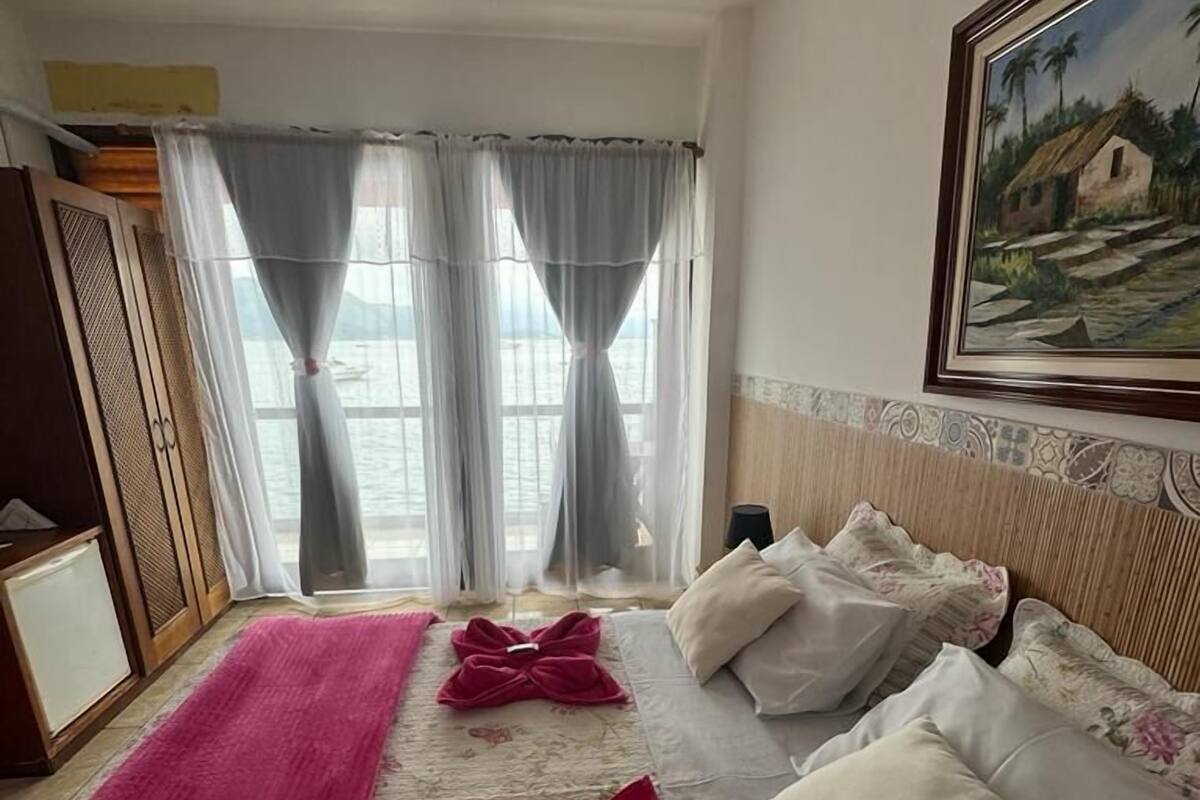 A hotel room with a bed, a large window with curtains, a painting on the wall, and a red rug.