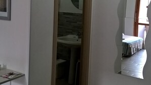 A narrow hallway with a wooden door leading to a bathroom with a sink and mirror. There is a small window with a curtain and a trash bin on the left. - Vacanza a Casa (Rome)