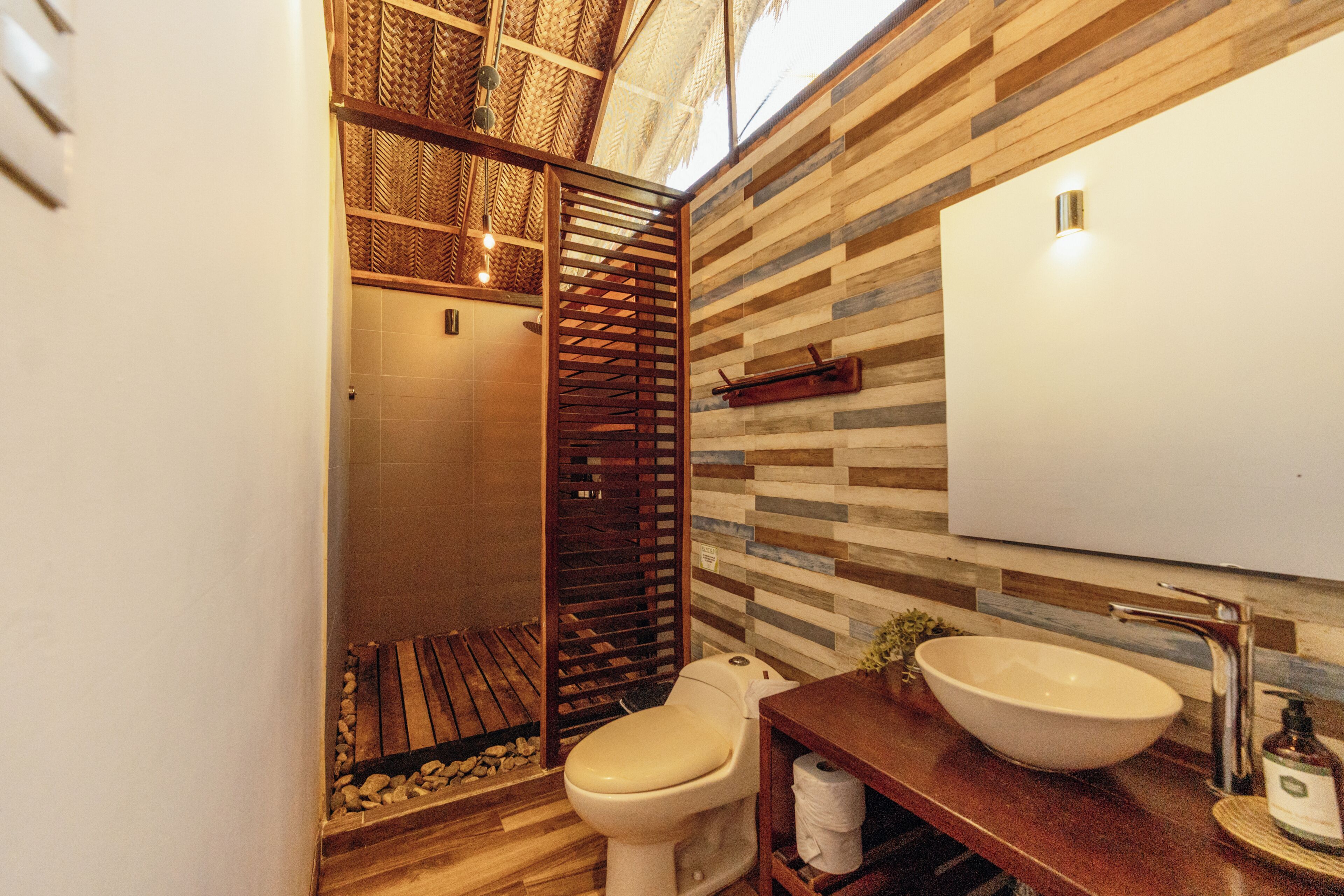 Un baño con suelo de madera, inodoro blanco, cabina de ducha de madera y un espejo grande.