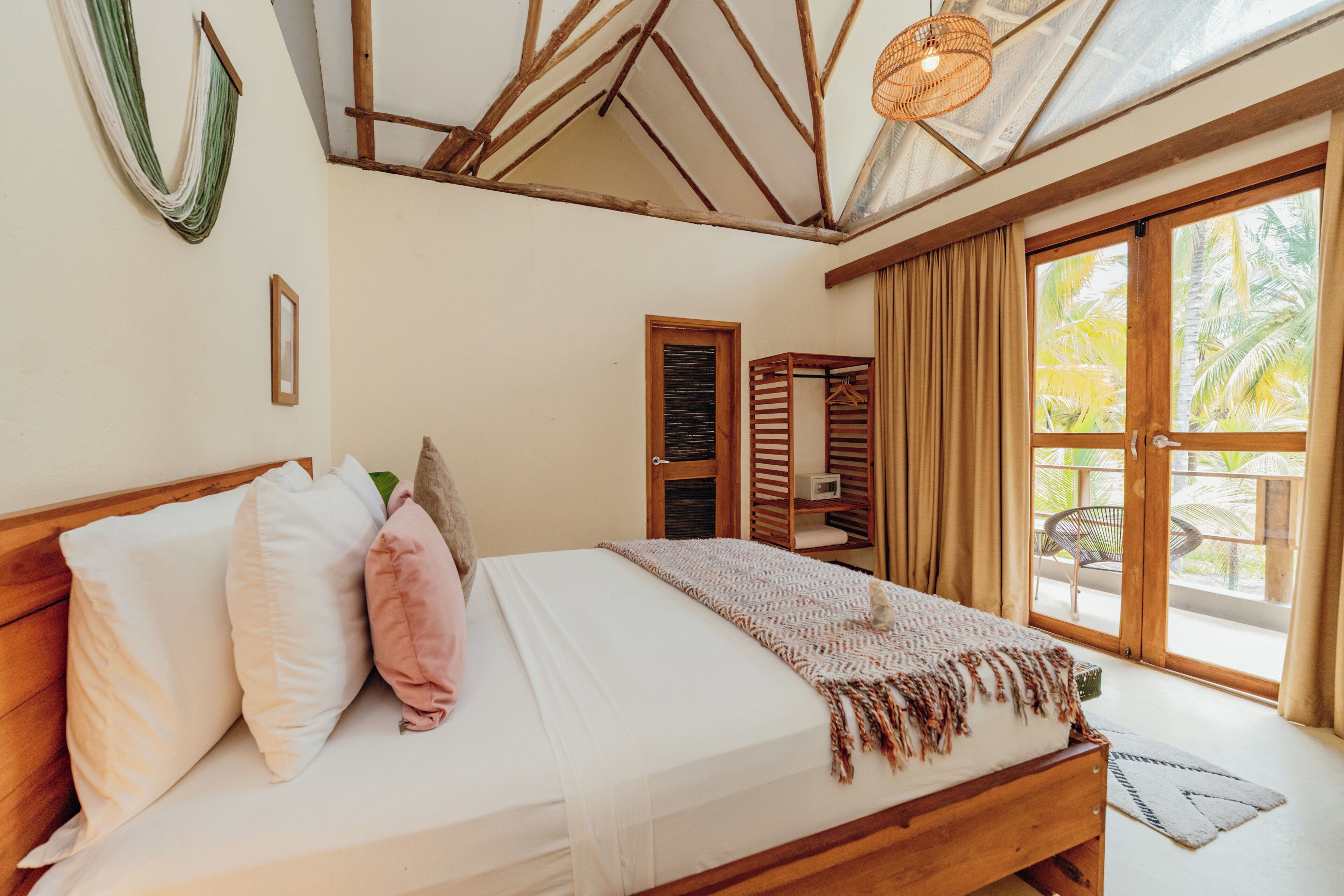 A bedroom with a wooden bed, a hanging light fixture, a window with curtains, and a view of palm trees.