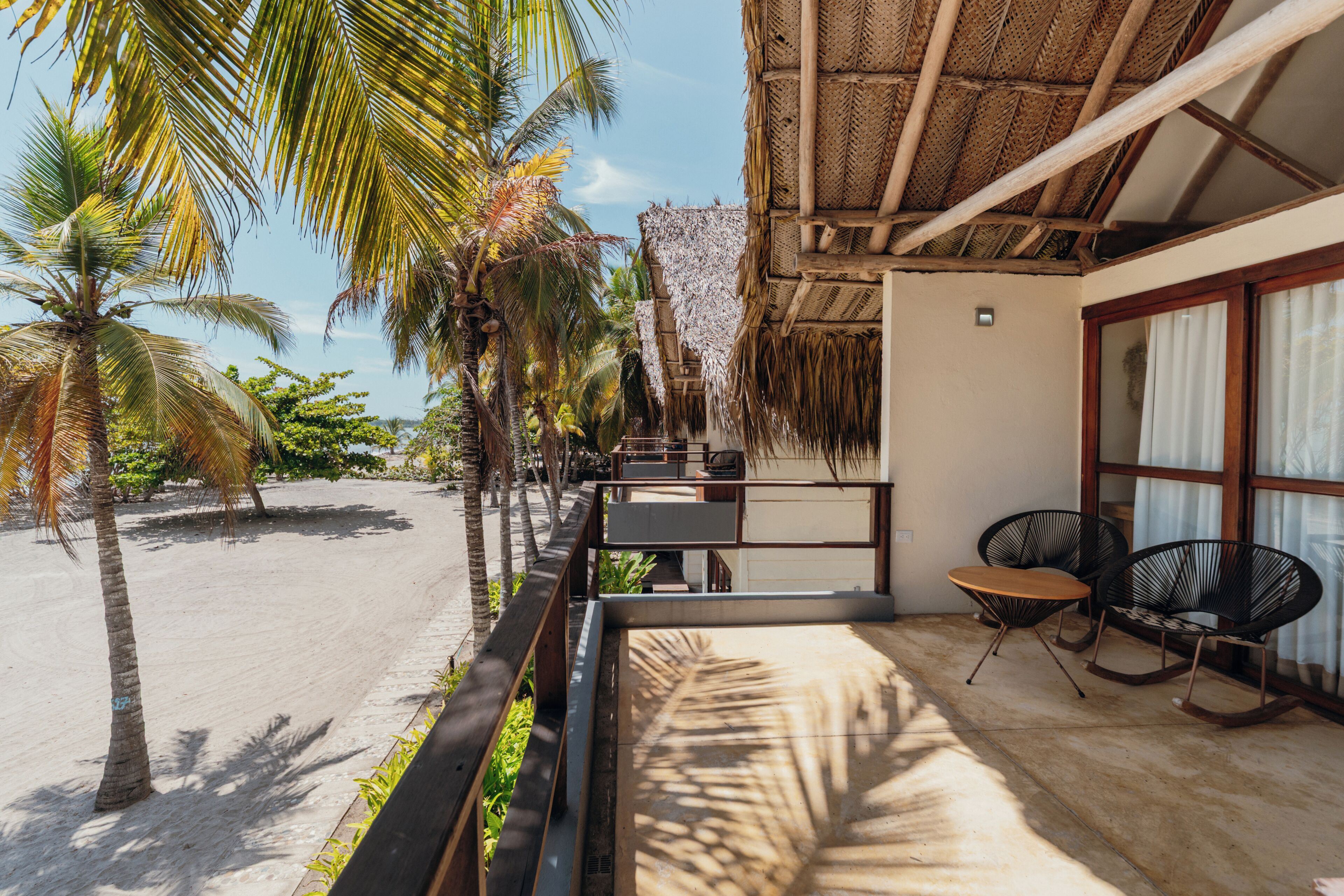 A beachside balcony with a thatched roof, overlooking a sandy beach and palm trees.