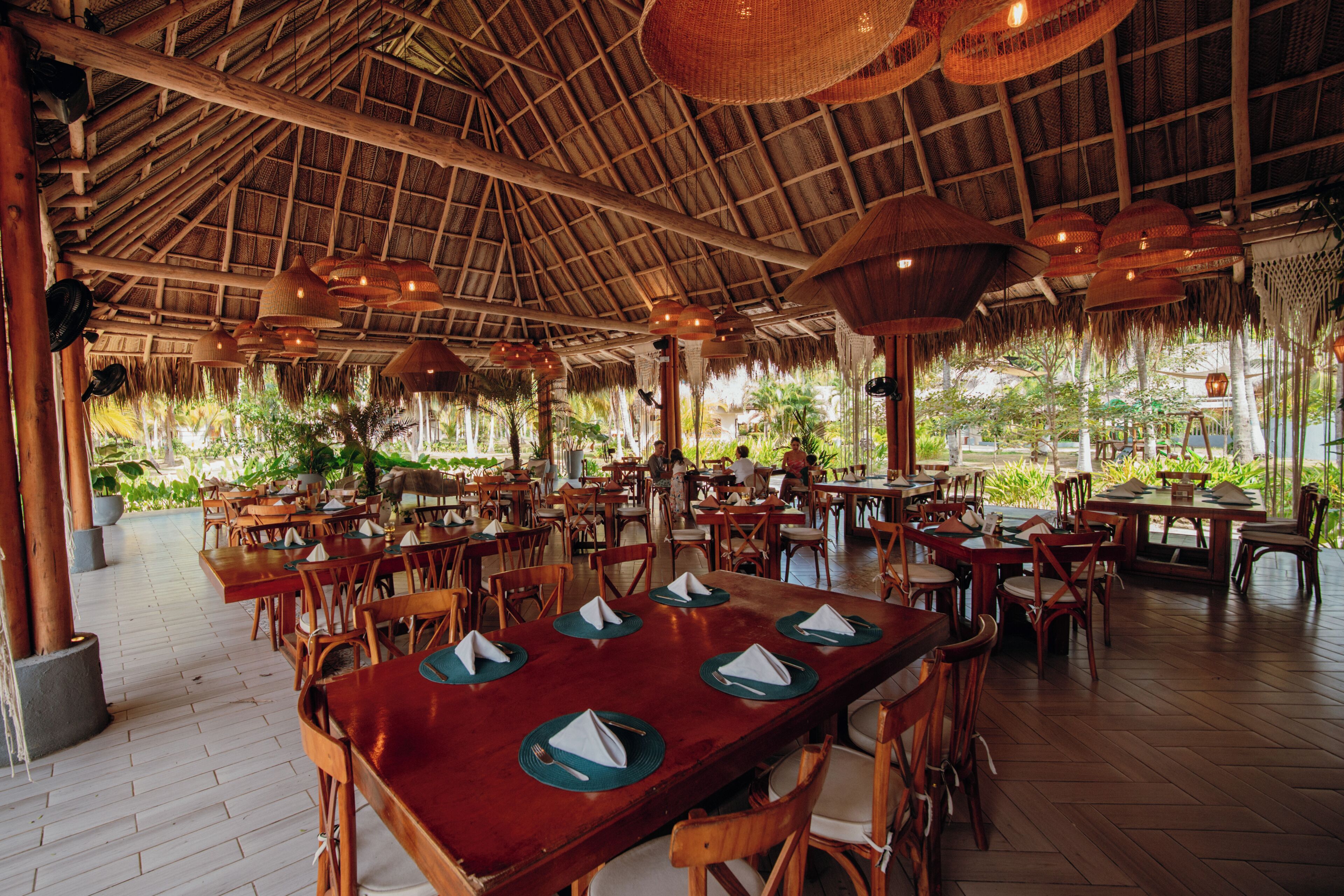 A spacious outdoor dining area with wooden tables and chairs, a thatched roof, and potted plants.