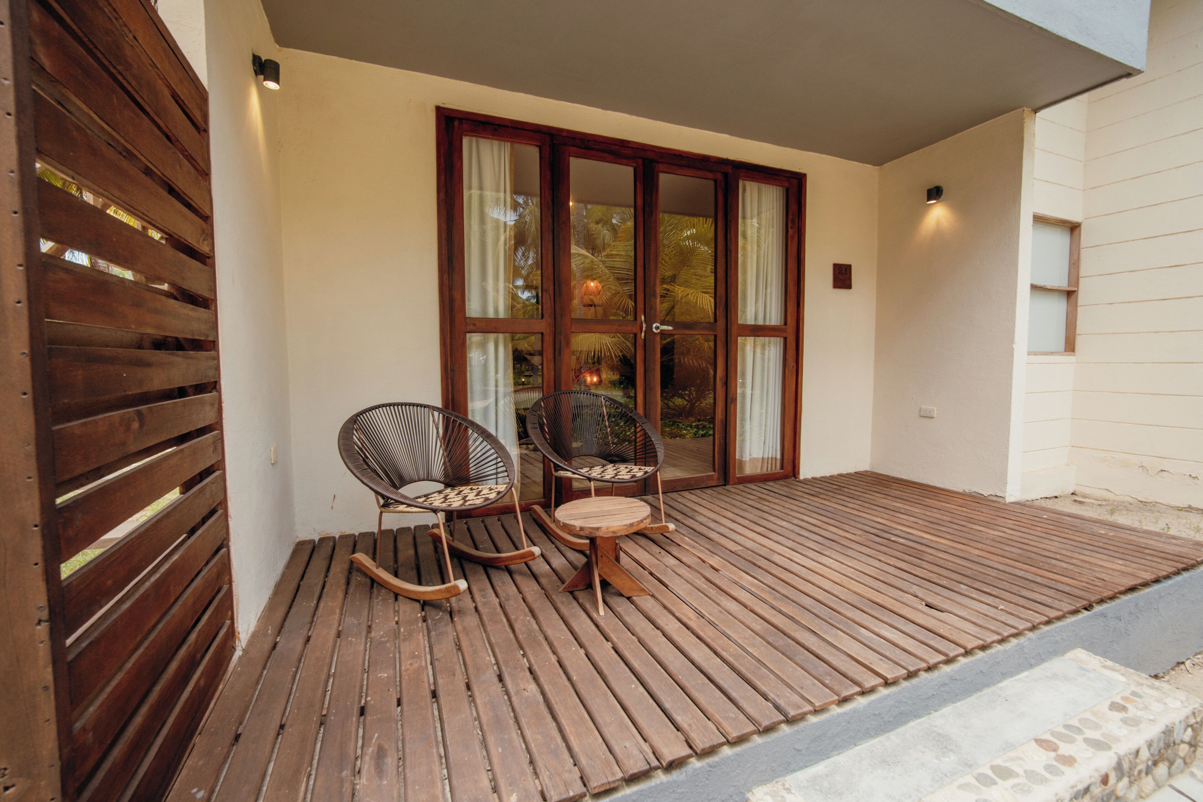 Terraza de madera con dos mecedoras y una mesita, que conduce a una puerta de cristal con cortinas.