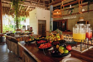 Un bufé con frutas y bebidas variadas, muebles de madera y plantas colgantes.