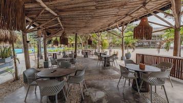Een terras buiten met tafels en stoelen, een rieten dak en uitzicht op het zwembad.