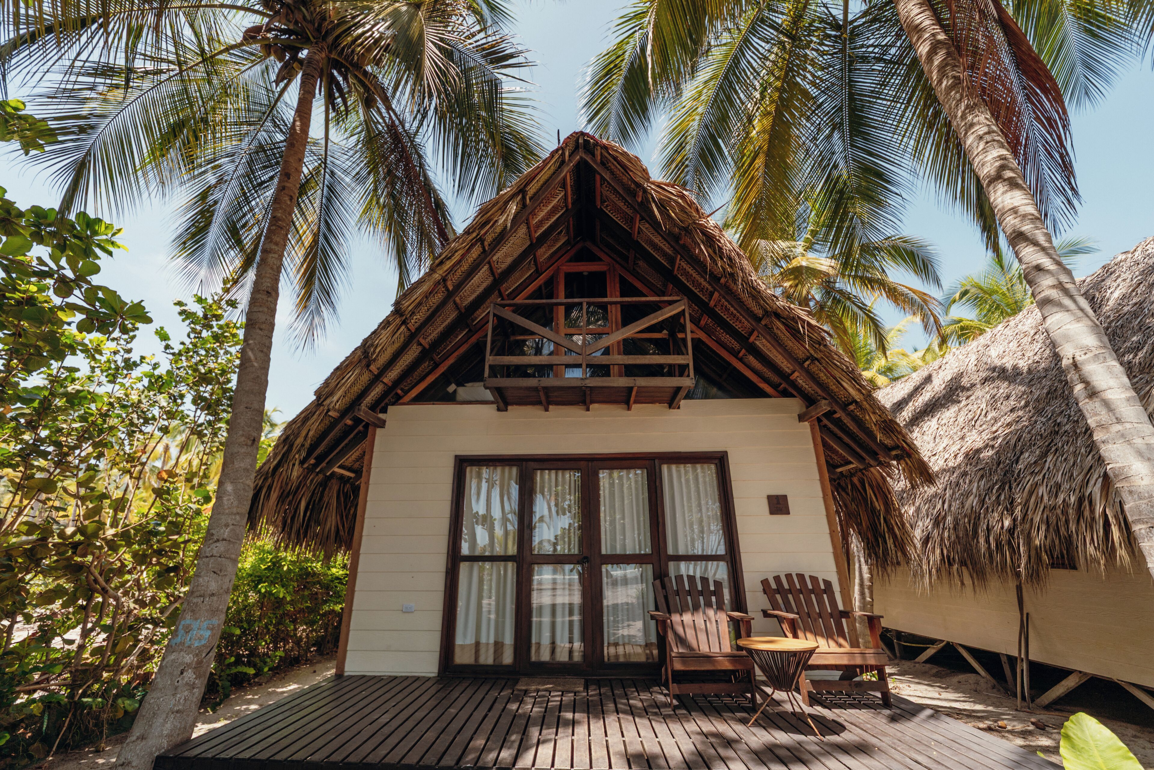 A thatched-roof bungalow with a wooden deck, two wooden chairs, and a large window.
