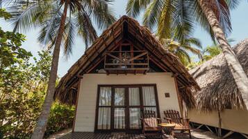 A thatched-roof bungalow with a wooden deck, two wooden chairs, and a large window.
