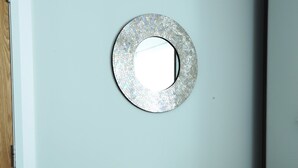 Ein Zimmer mit hellblauer Wand, einem runden Spiegel mit Mosaikrahmen und einem cremefarbenen Sessel.