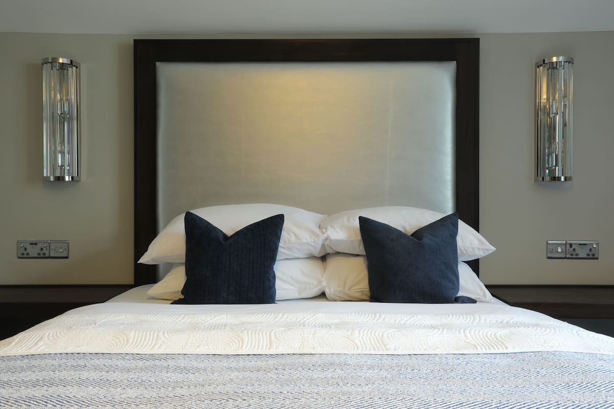 Una cama bien tendida con una colcha blanca y azul estampada, dos almohadas oscuras y un cabecero grande. Hay dos lámparas de pared a cada lado de la cama.