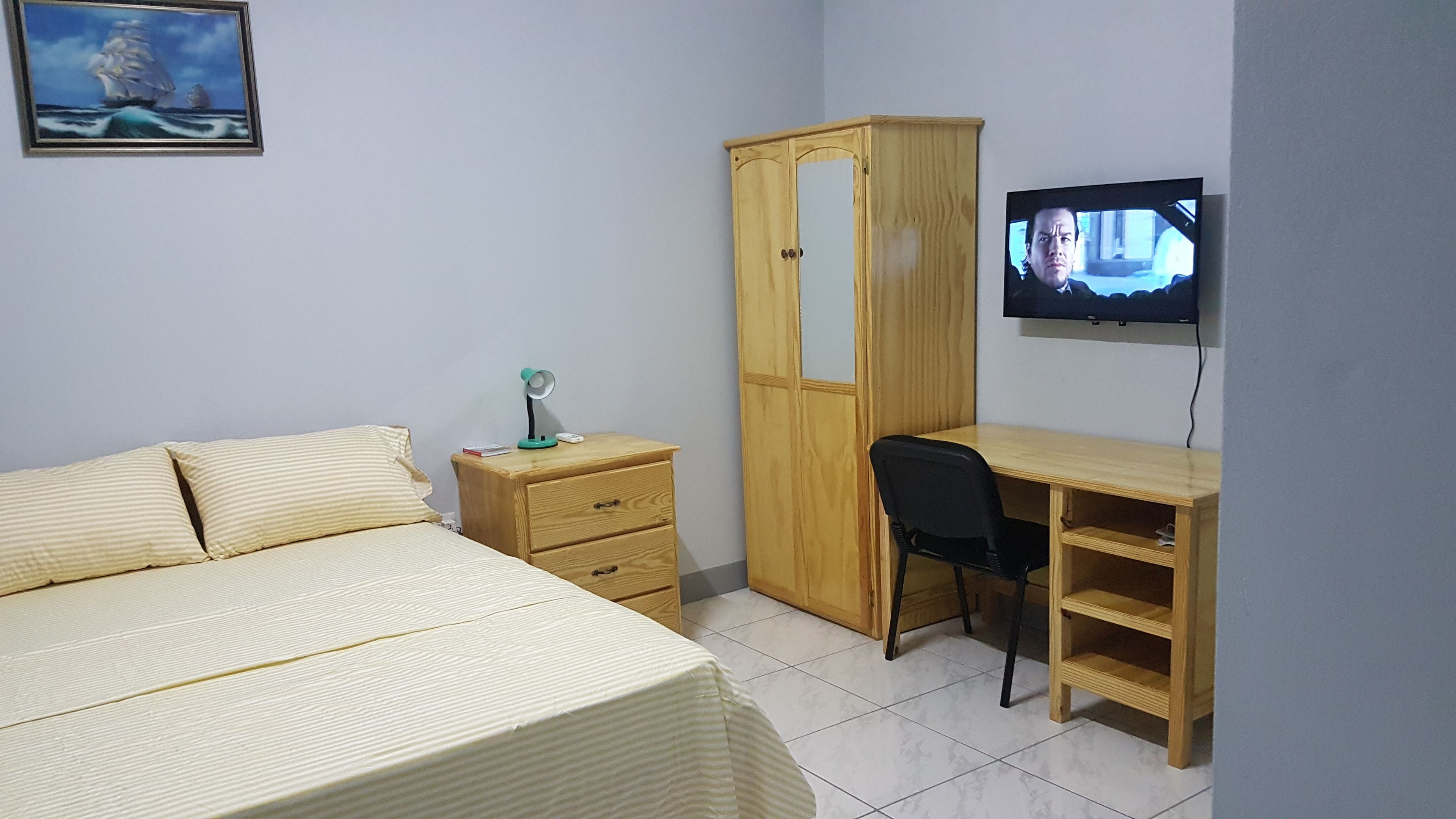 A bedroom with a bed, a wooden wardrobe, a desk with a chair, and a television on the wall.