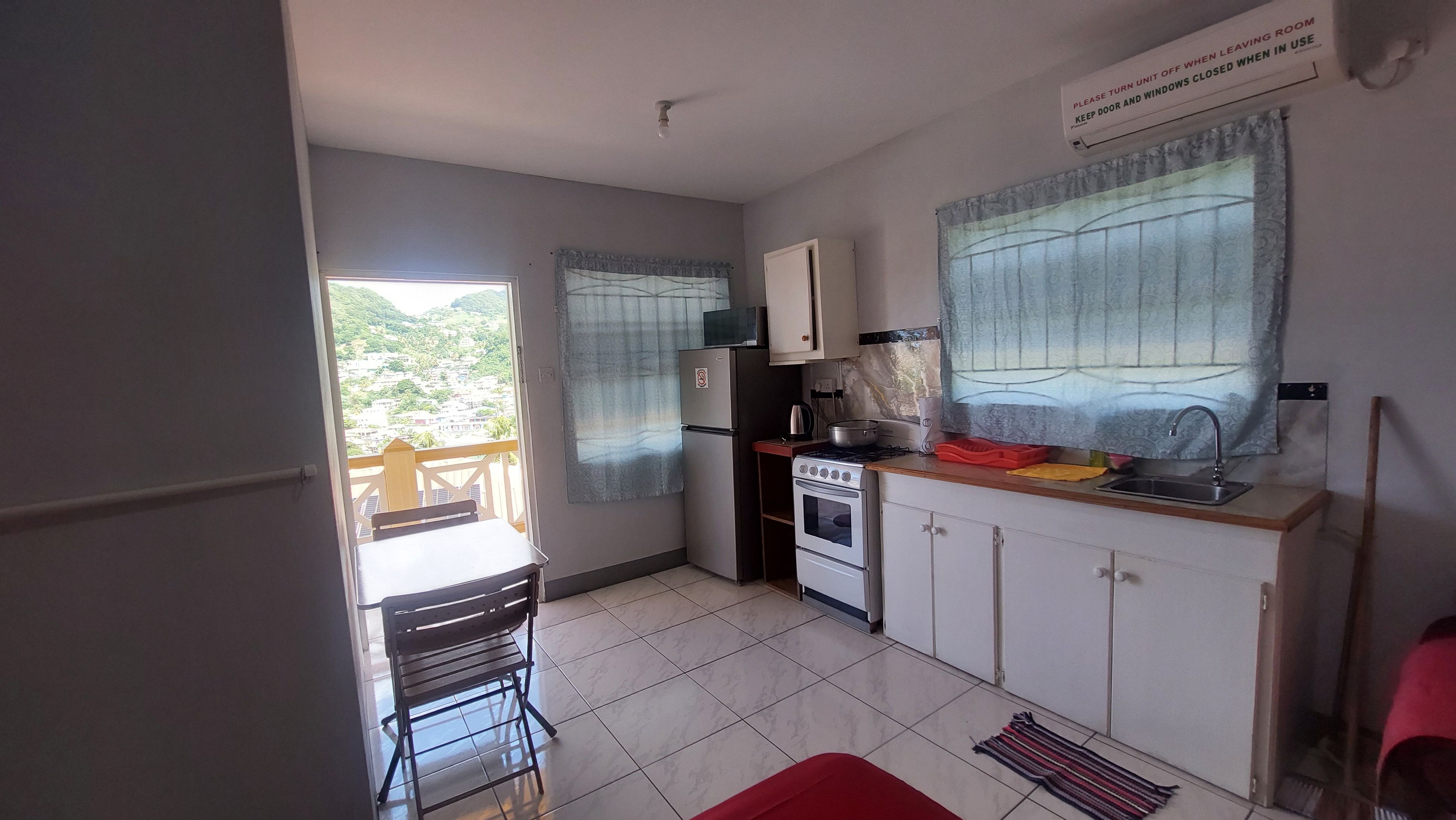 A compact kitchen with a refrigerator, stove, microwave, and sink. A window with curtains allows natural light, and a view of a green landscape is visible.
