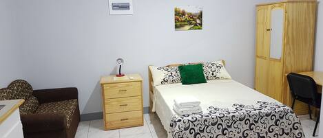 A bedroom with a bed, a wooden wardrobe, a desk, a chair, a small wooden nightstand with a lamp, and a picture on the wall.