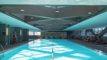 Uma piscina interior espaçosa com água azul cristalina, rodeada por espreguiçadeiras e cadeiras de banho.