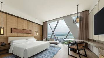 A hotel room with a large bed, a desk, a chair, a TV, and a view of a cityscape.