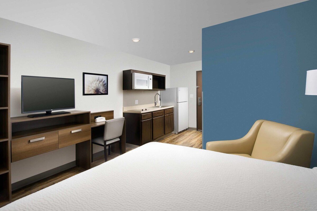 A hotel room with a bed, a desk with a computer, a chair, a television, a kitchen area with a refrigerator, and a blue accent wall.