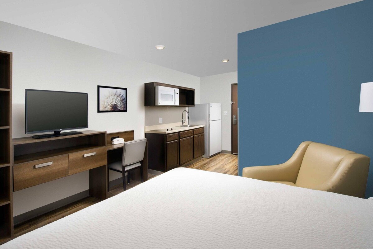 A modern hotel room with a bed, a desk with a computer, a chair, a television, a kitchen area with a microwave, and a sink.