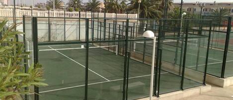 Una cancha de tenis al aire libre con superficie verde, rodeada por una valla metálica y palmeras.